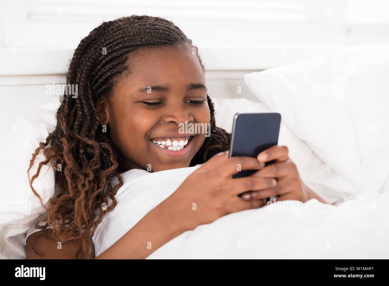 Afrikanische Mädchen liegt auf dem Bett unter der Decke mit Handy Stockfoto