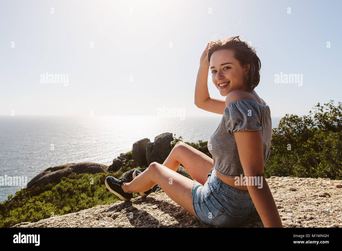 Lächelnde Frau sitzt auf dem Gipfel des Berges und genießen herrliche Natur. Junge weibliche am Berg mit wunderschönem Blick aufs Meer. Stockfoto