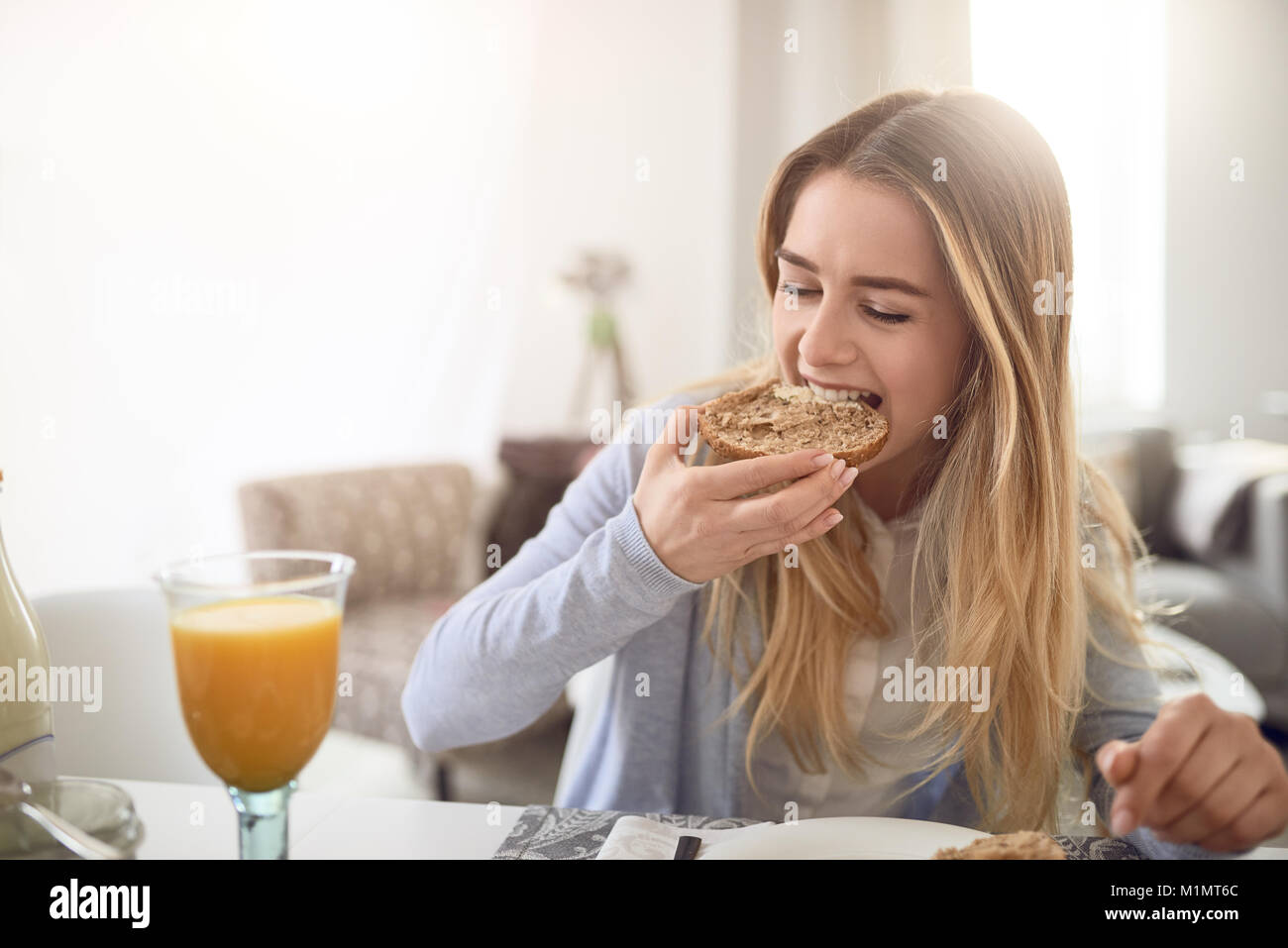 Hübsche junge jugendmädchen einen Bissen von einem gesunden braunen Vollkorn rollen, wie Sie ein gesundes Frühstück zu Hause genießt bei einem Glas frischen Orange juic Stockfoto