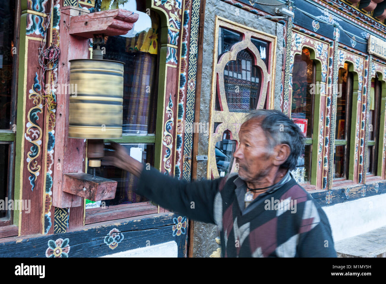 Jakar, Bumthang, Bhutan. Mann Drehen einer Gebetsmühle, als er von einem Store Eingang geht. Stockfoto