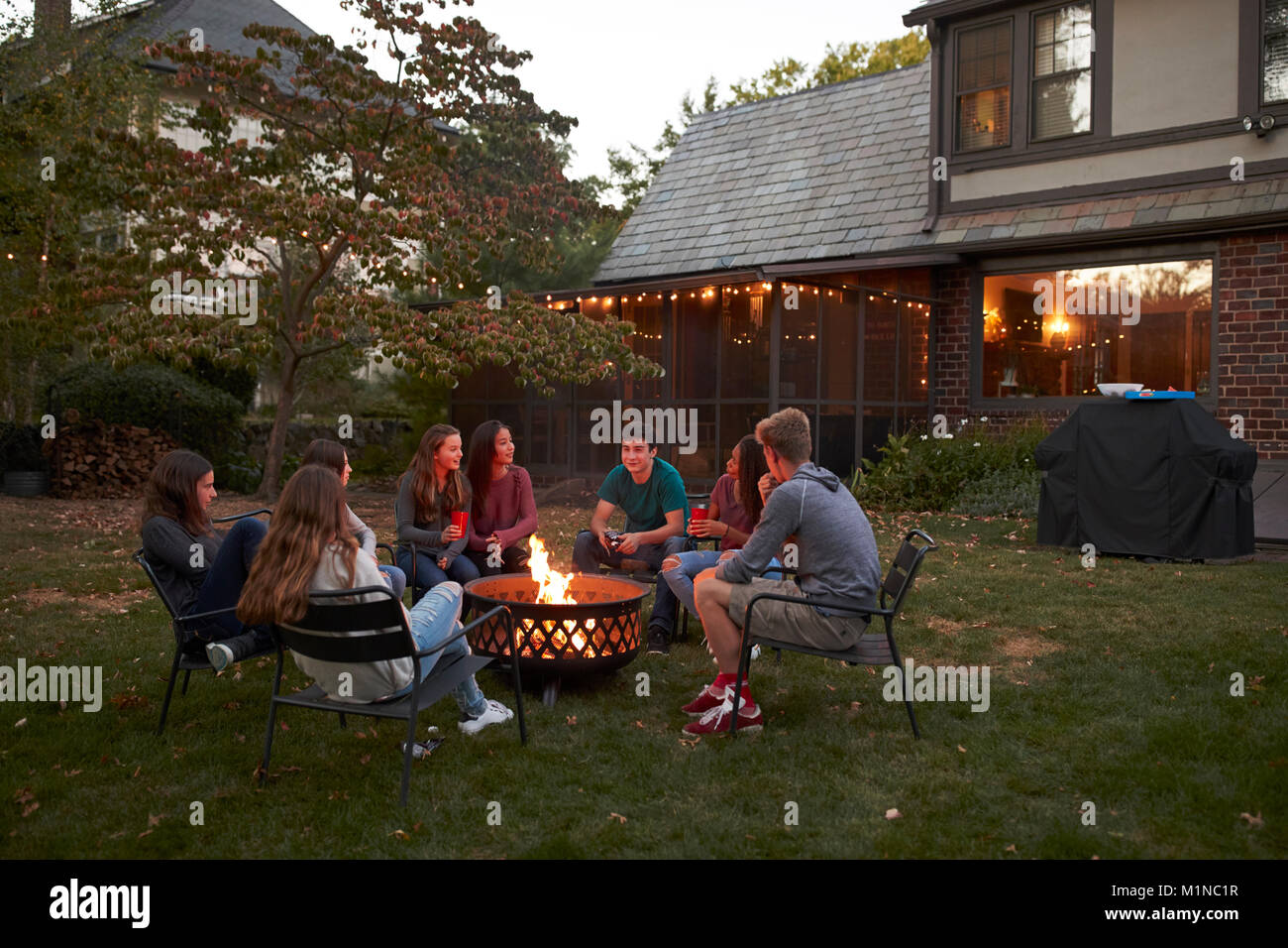 Jugendliche sitzen Reden um eine Feuerstelle im Garten bei Dämmerung Stockfoto