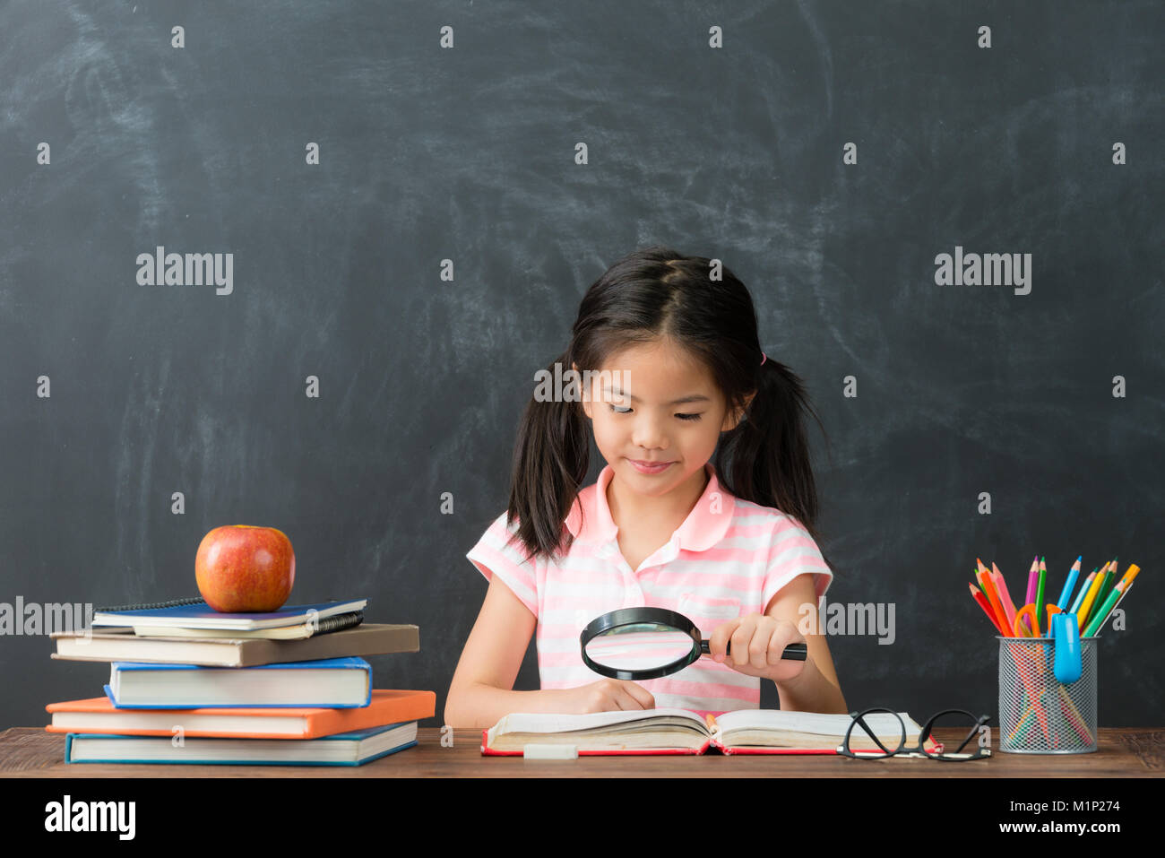 Attraktive süße kleine Mädchen mit Lupe Tool lesen Buchen Sie im blackboard Hintergrund und Erforschung Schule Hausaufgaben zu studieren. Stockfoto