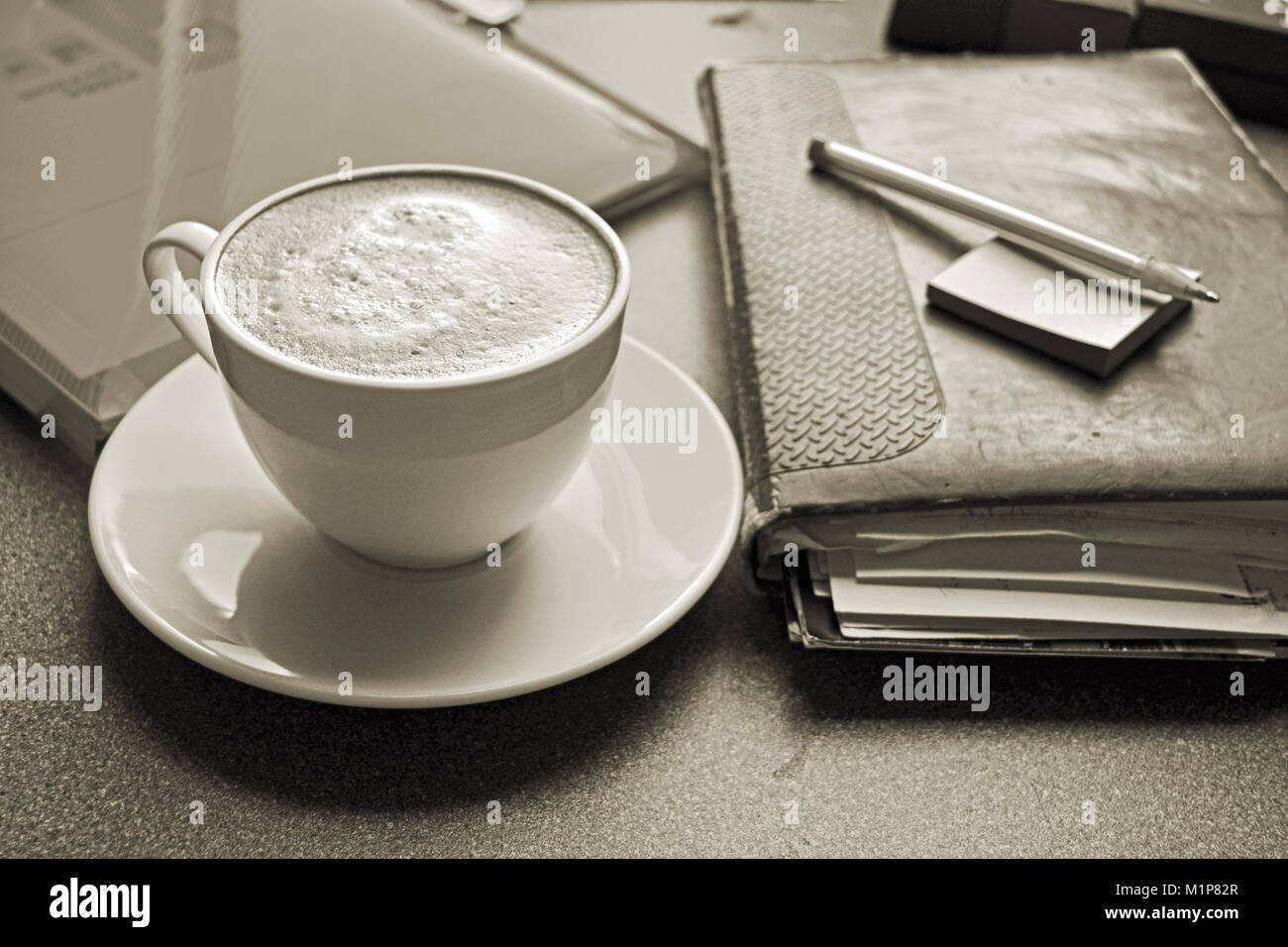 Bei einer Tasse frisch gebrühtem Kaffee auf einem Schreibtisch mit einem Tagebuch und Stift, in Sepia behandelt Stockfoto