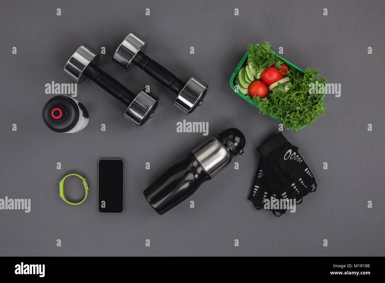 Flach mit Smartphone mit Fitness Tracker, Hanteln und Salat isoliert auf Grau Stockfoto