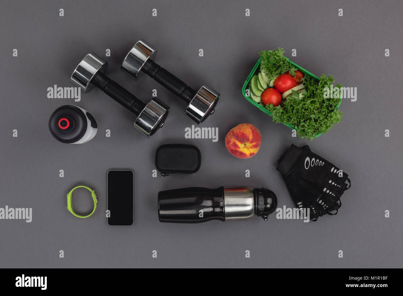 Draufsicht auf das Smartphone mit Fitness Tracker, Hanteln und Salat isoliert auf Grau Stockfoto