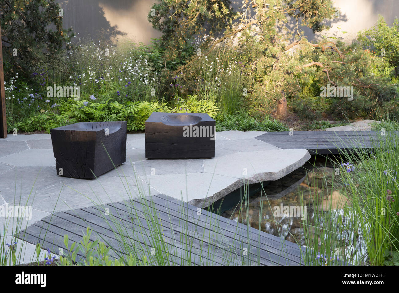 Moderner Garten mit gepflastertem Innenhof mit Holzterrasse und kleinem Teich Chelsea Flower Show UK Holzsteg über dem Pool, verkohlter Holzwürfelsitz Stockfoto
