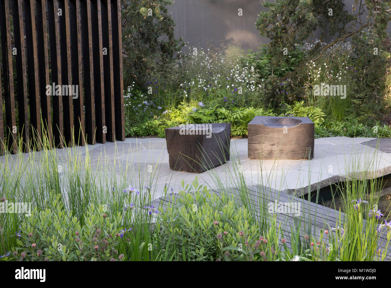 Kleiner Garten mit gepflasterter Terrasse und modernen Holzgartenmöbeln im Freien dunkler Holzgang über Teich verkohlter Holzwürfelsitz UK Stockfoto