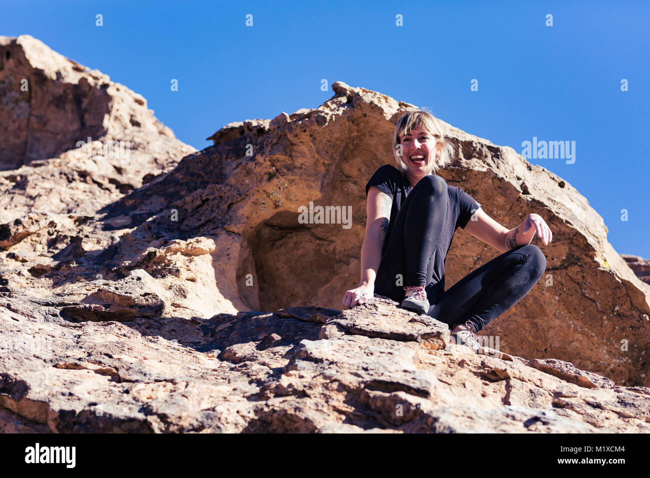 Dünne blonde kaukasische Frau mit Arm Tattoos sitzt triumphierend nach Klettern am Felsen in der Wüste von Kalifornien Stockfoto