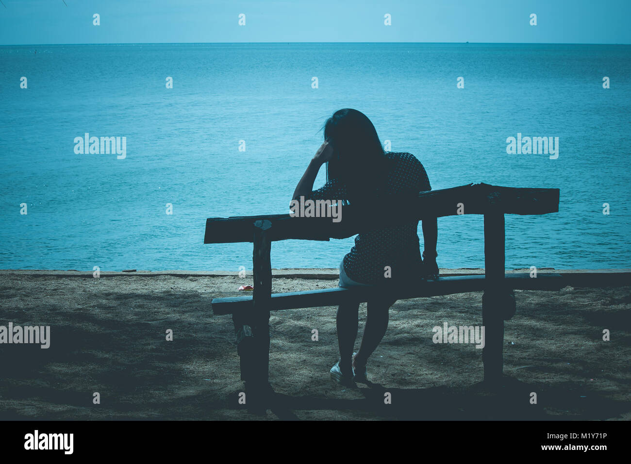 Silhouette der einsamen Frau sitzt auf der Holzbank am Strand und auf der Suche nach blauen Meer am Abend. Stockfoto