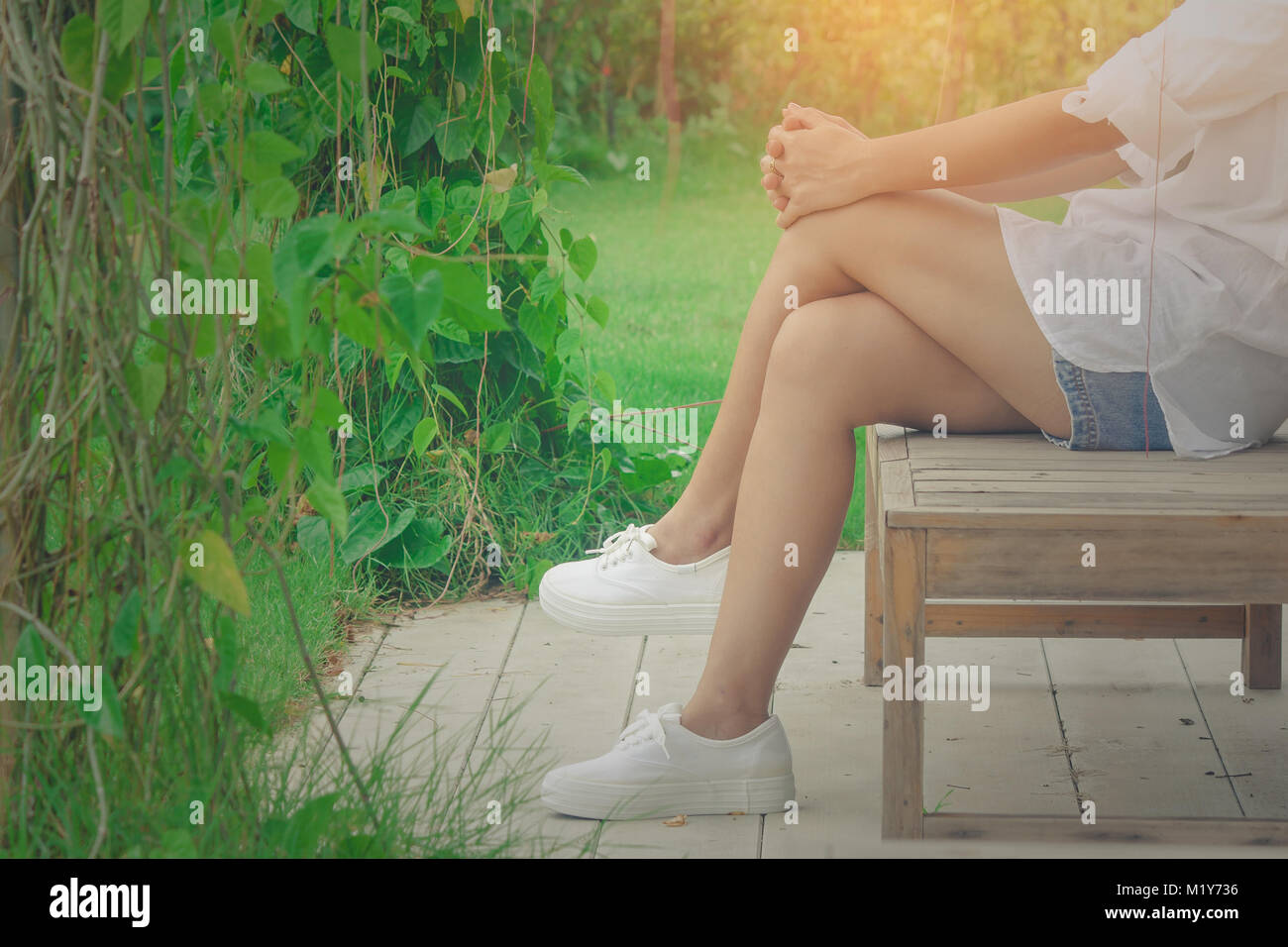 Entspannung Konzept: Frau trägt ein weisses T-Shirt und kurze Jean, ihre Entspannung auf hölzernen Stuhl am Garten mit grünen Hintergrund umgeben. Stockfoto