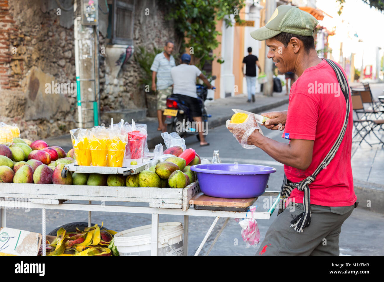 Cartagena, Kolumbien - Januar 23th, 2018: eine Frucht street Hersteller schneiden Mango stehen auf der Straße mit seinem Holz- Warenkorb mit Früchten in Cartagena. Stockfoto