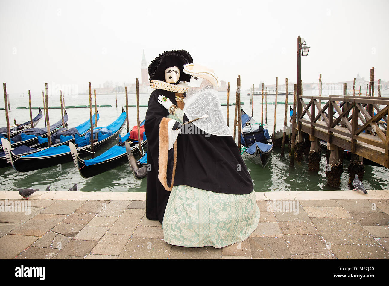 Karneval in Venedig - Paar in der Liebe der Venezianischen Masken auf den Markusplatz in Venedig mit Gondeln im Hintergrund. Stockfoto