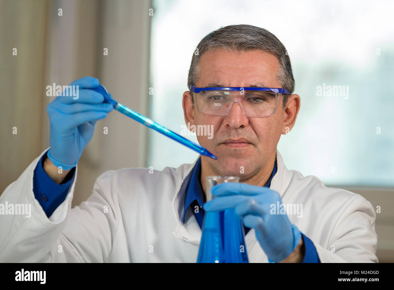 Forscher trägt blaue, weiße Uniform Schutzhandschuhe und Schutzbrille. Pipette Drop blau Probe in einen Becher. Stockfoto