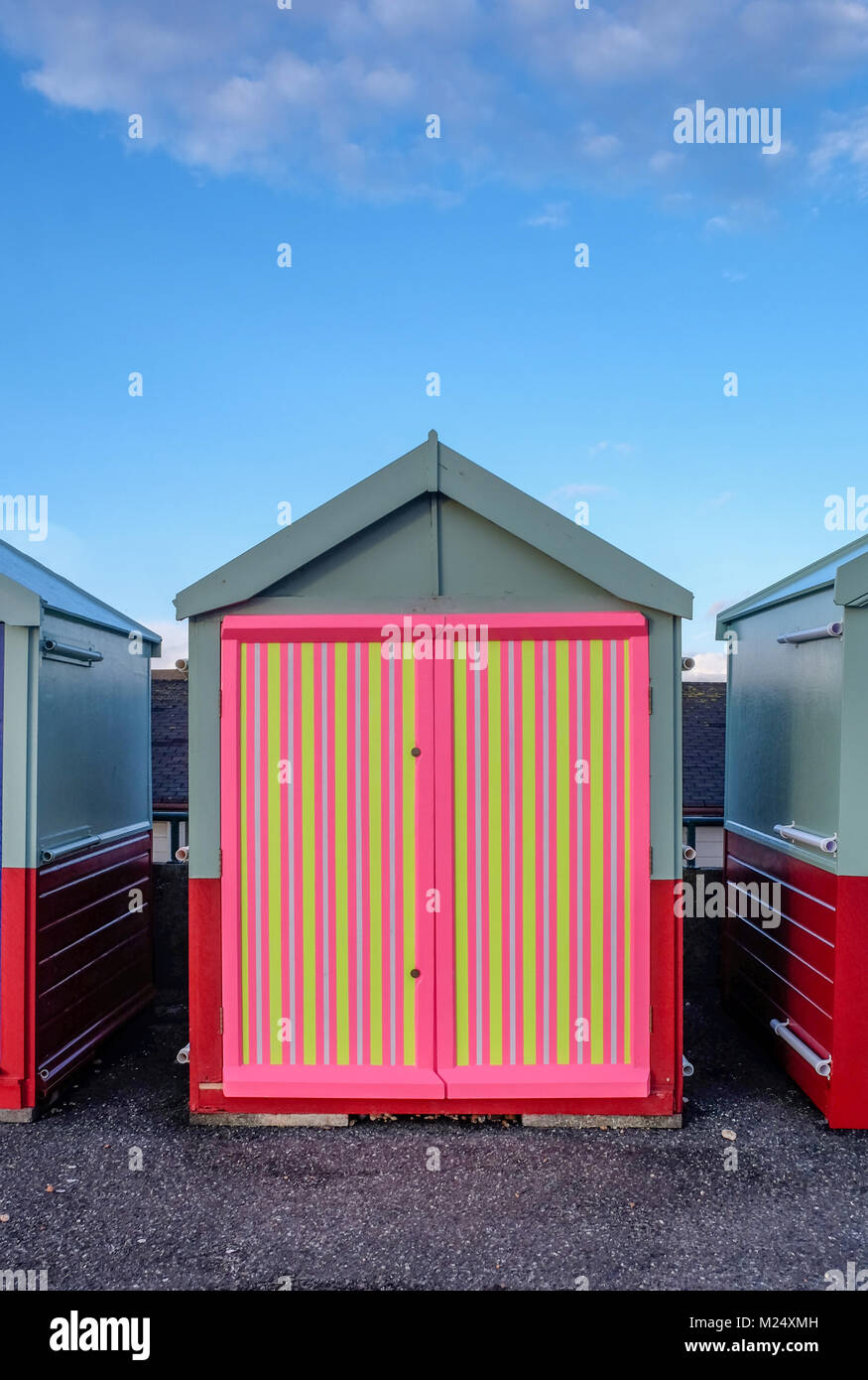 Brighton Beach Hütte mit einem bunten Tür von Gelb, Pink, Schwarz Streifen hinter ist blauer Himmel und Infront ist die konkrete Promenade. Stockfoto