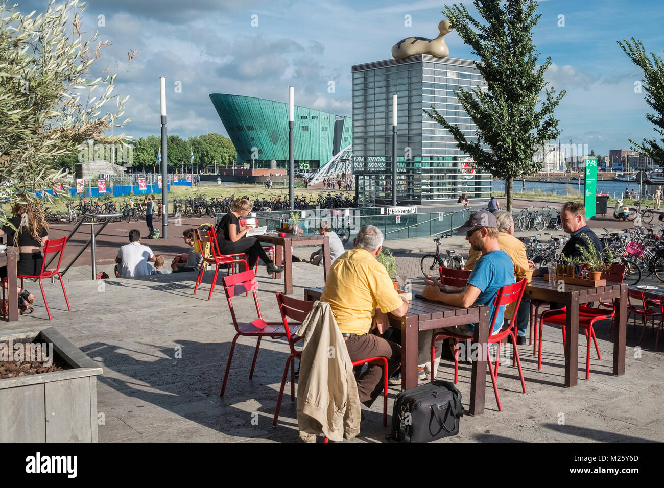 Die Menschen in der Sonne außerhalb der öffentlichen Bibliothek von Amsterdam sitzend, mit NEMO Science Museum im Hintergrund, Oosterdokskade, Amsterdam, Niederlande Stockfoto