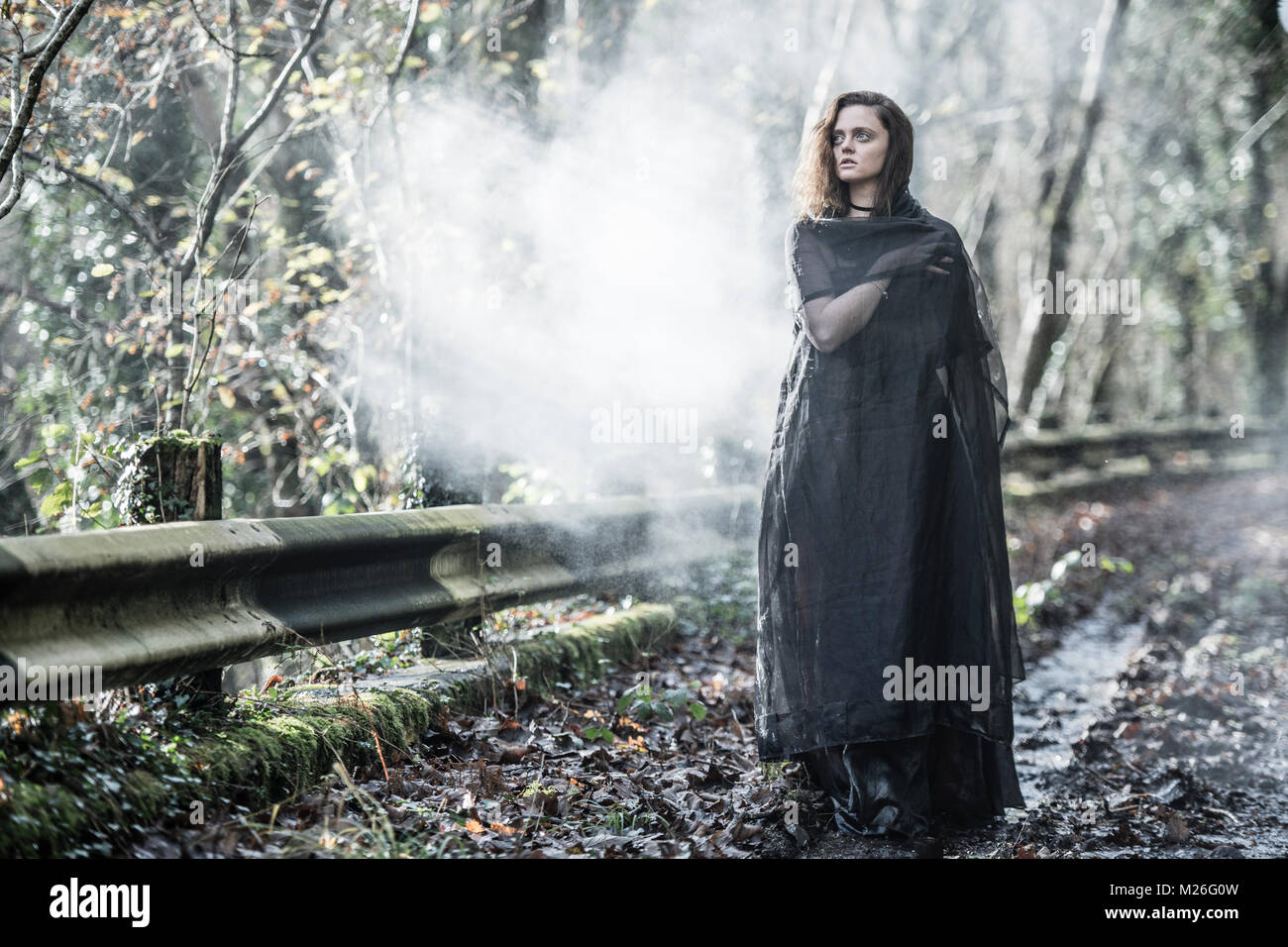 Frau von Mystery/Goth: eine junge Frau in einem schwarzen Kleid und Umhang gekleidet, allein auf einem Nebelhaften verlassenen Straße Stockfoto
