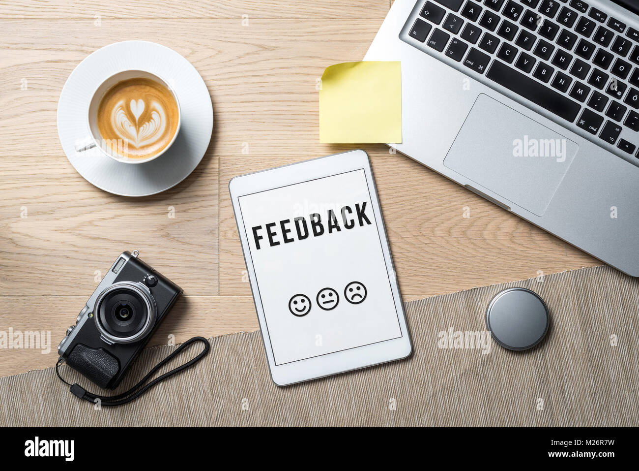 Feedback Schreiben mit Emoticons auf Tablet Bildschirm mit Kamera, Kaffeebecher und Laptop liegen auf Büro Schreibtisch als Flach Stockfoto
