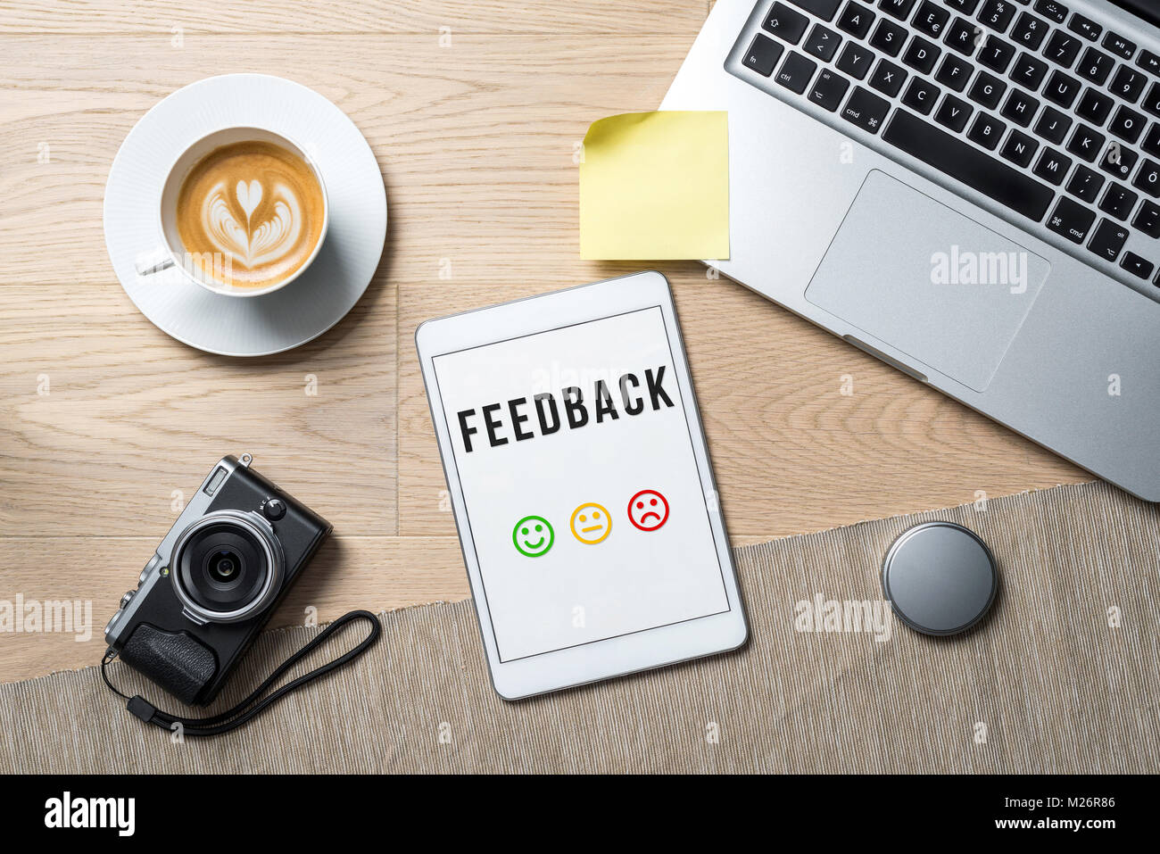 Feedback Schreiben mit bunten Emoticons auf Tablet Bildschirm mit Kamera, Kaffeebecher und Laptop liegen auf Büro Schreibtisch als Flach Stockfoto
