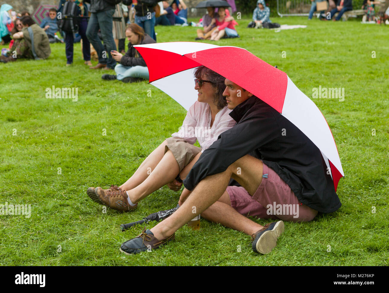 Ein paar in Shorts Schutz vom britischen Sommer Wetter unter einem Sonnenschirm sitzend auf dem Gras zu einem Outdoor Event. Stockfoto