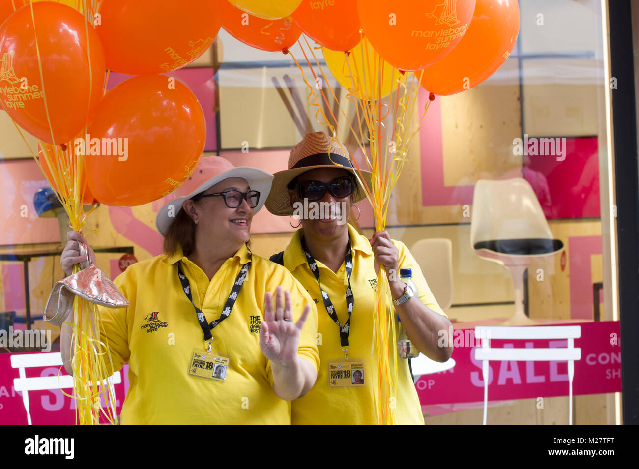 Zwei Freiwillige lächelnd in den gelben Hemden Holding orange Luftballons an der Marlyebone Sommer Fayre Marlyebone Food Festival in London an einem sonnigen Tag Stockfoto