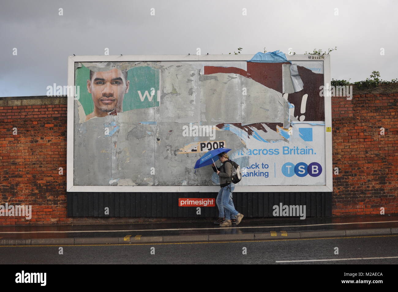 Zwei Menschen zu Fuß mit Sonnenschirmen im Regen vor einem Werbung Plakatwand in Bristol, Großbritannien Stockfoto