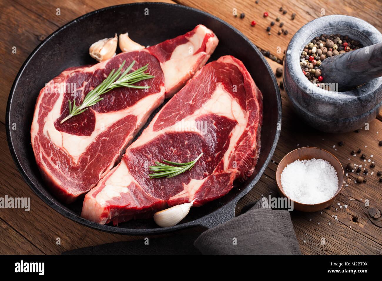 Zwei frische rohe Marmor Fleisch, Black Angus ribeye Steak mit Gewürzen in eiserne Pfanne auf einem alten rustikalen Tisch. Rohes Rindfleisch auf einer hölzernen Hintergrund. Ansicht von oben Stockfoto