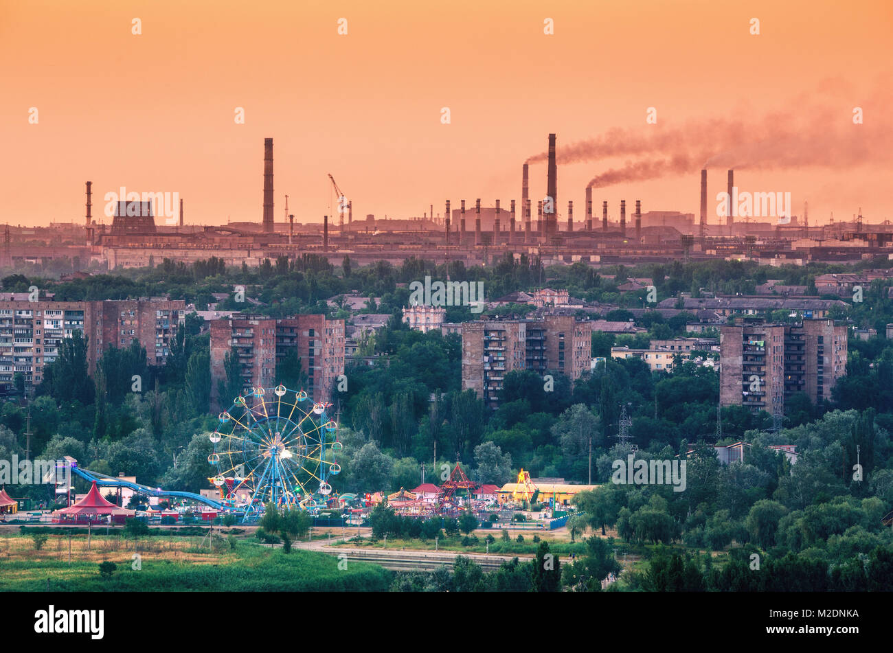 Vergnügungspark mit Riesenrad und Gebäude auf dem Hintergrund der Metallurgie Anlage mit Smog bei Sonnenuntergang. Ökologie Probleme, Luftschadstoffe. Pi Stockfoto