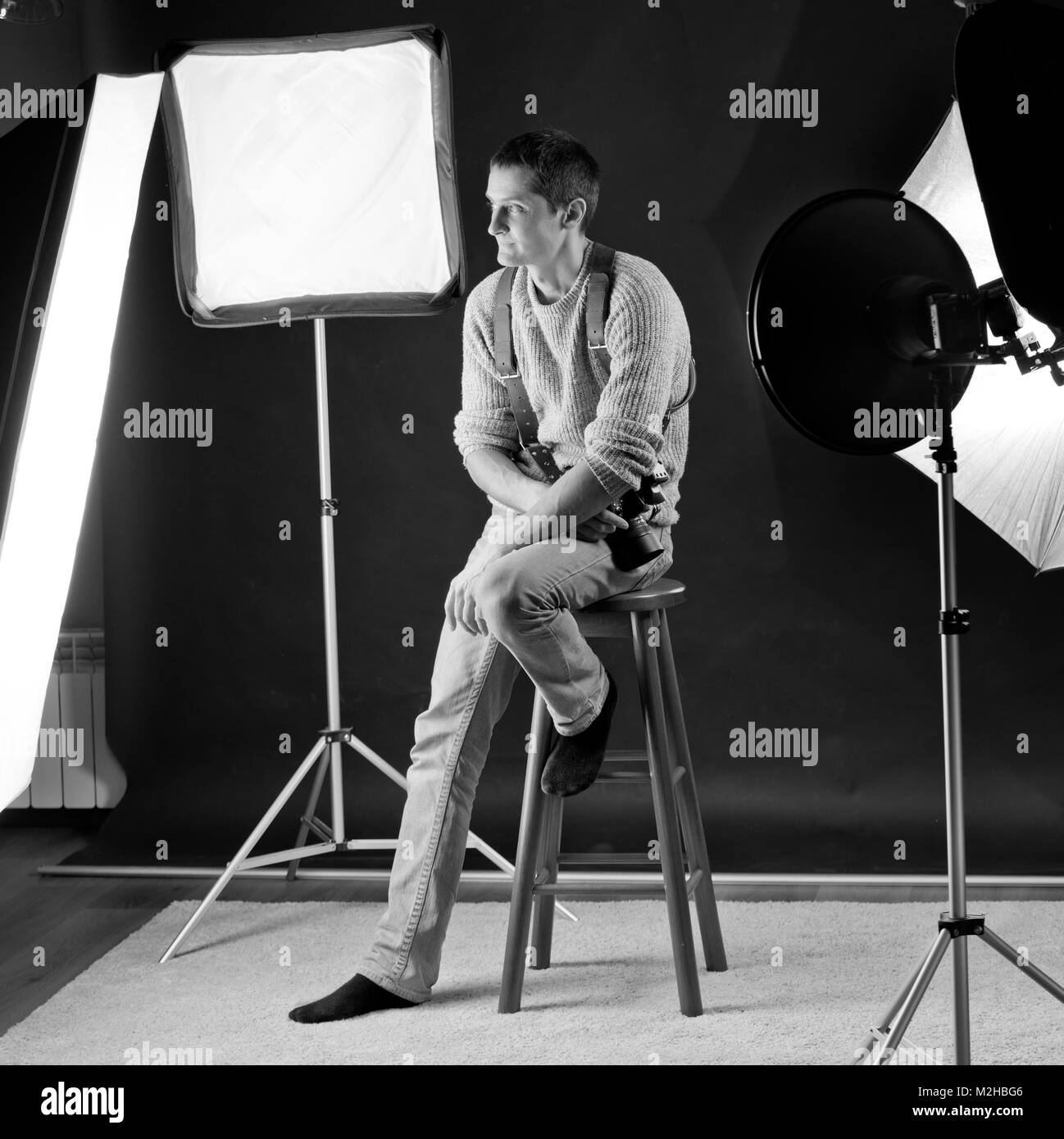 Fotograf im Studio mit seinem Licht Modifikatoren um und saß auf einem Bar Stuhl, schwarzen Hintergrund. Er wird von Softboxen, Beauty Dish, Regenschirm umgeben Stockfoto