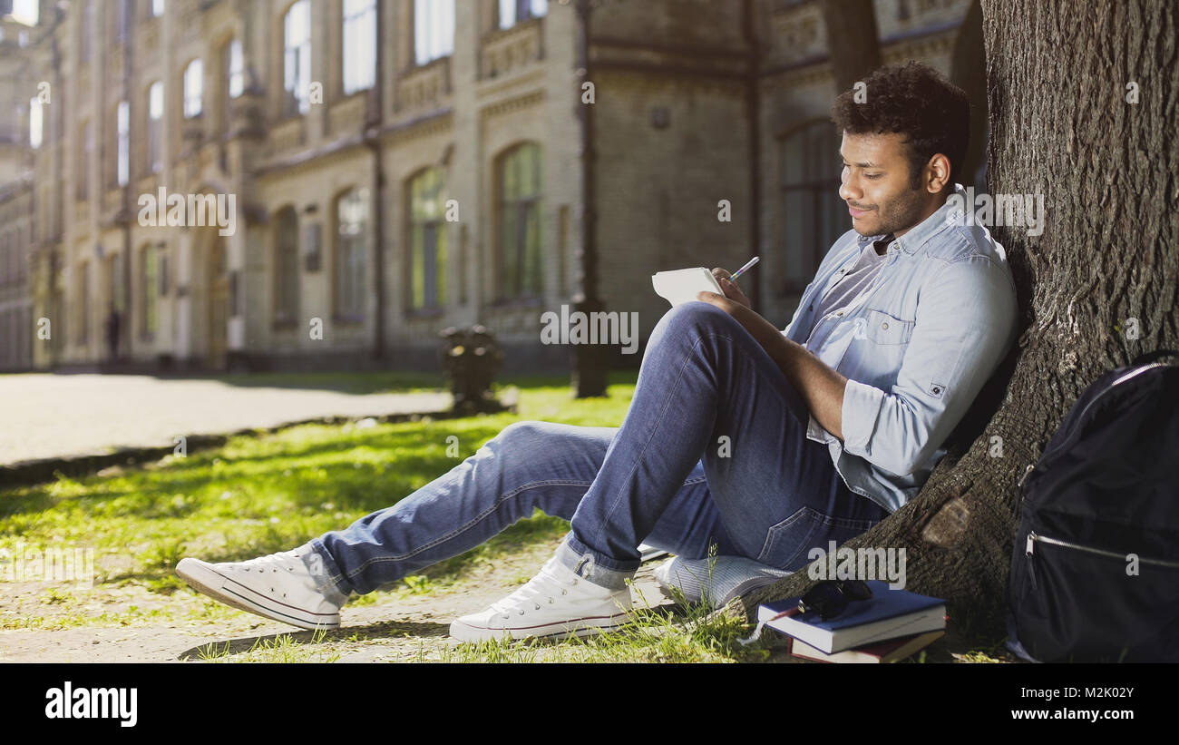 Mixed-race jungen Kerl sitzen unter Bäumen, Notizen im Notebook, inspiration Stockfoto