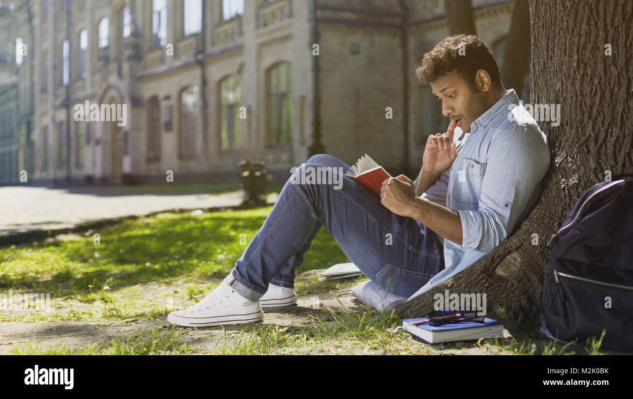 Multirassischen jungen Kerl sitzen unter Bäumen, lesen interessantes Buch, bücherwurm Stockfoto