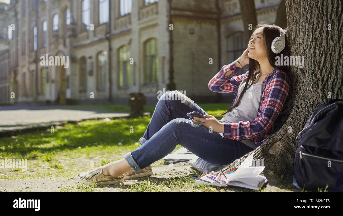 Weiblicher Kursteilnehmer sitzen auf Gras, lehnte sich gegen den Baum, Musik hören, entspannen Stockfoto