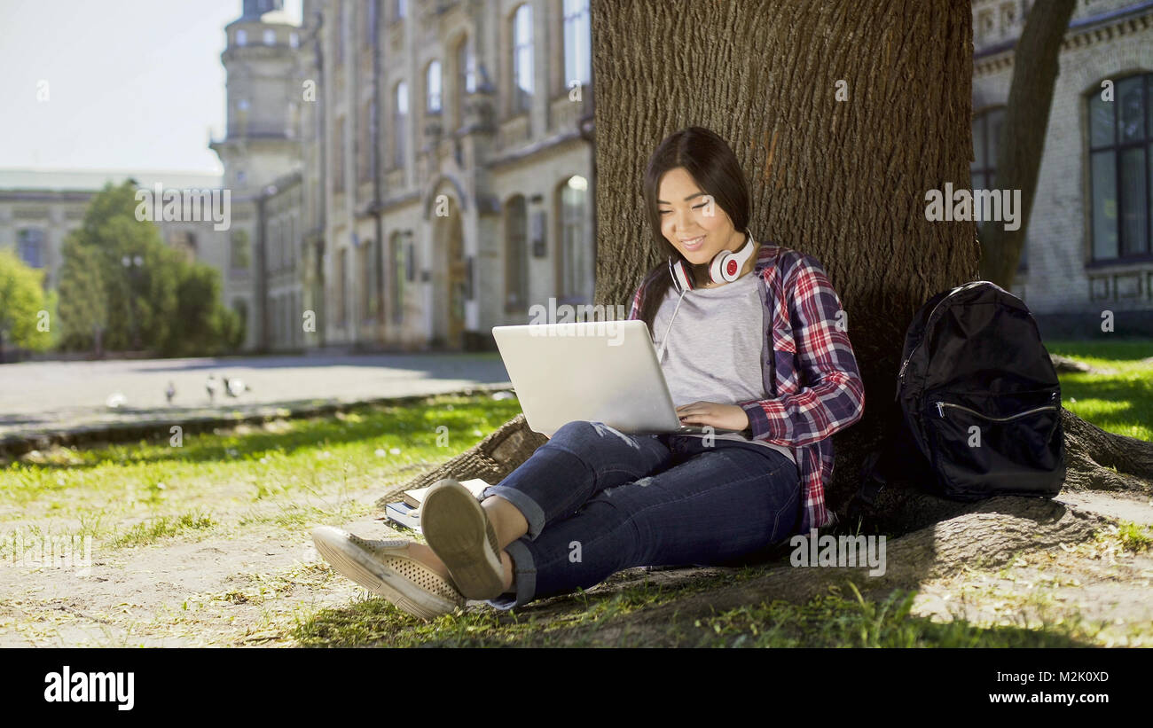 Studenten sitzen unter Baum in Campus, mit Laptop, Lächeln auf ihrem Gesicht Stockfoto
