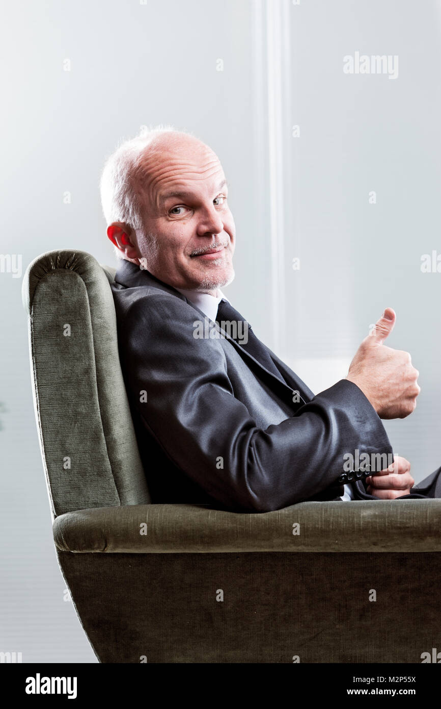 Happy Geschäftsmann einen Daumen nach oben Schild mit einem strahlenden Lächeln, als er in einem bequemen Sessel in der Nähe zu 7/8-Ansicht sitzt Zufrieden Stockfoto