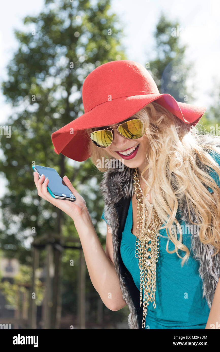 Junge Frau in der Nähe von Red Hat und Sonnenbrille Telefone außerhalb, semi-portrait Stockfoto