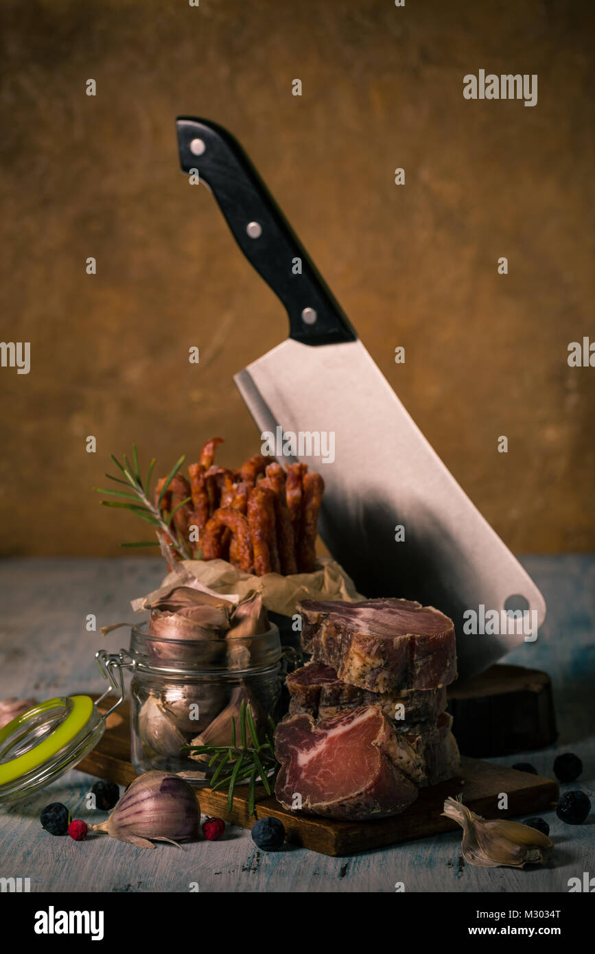 Vertikale Foto mit Raw getrocknet und geräuchert Toskana Fleisch von Wildschweinen. Das Fleisch ist zu große Stücke geschnitten und auf vintage Holzbrett mit Knoblauch in gestapelten Stockfoto