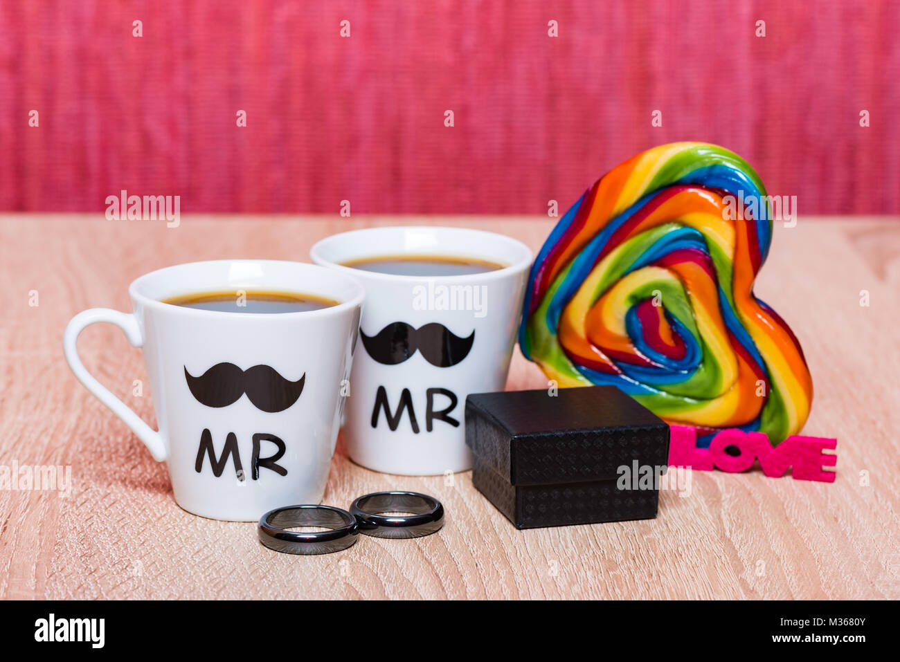 Homosexuelle Ehe Konzept. Zwei Tassen Kaffee, schwarzer Geschenkkarton mit Ehe Ringe und Rainbow lollipop Herz Stockfoto