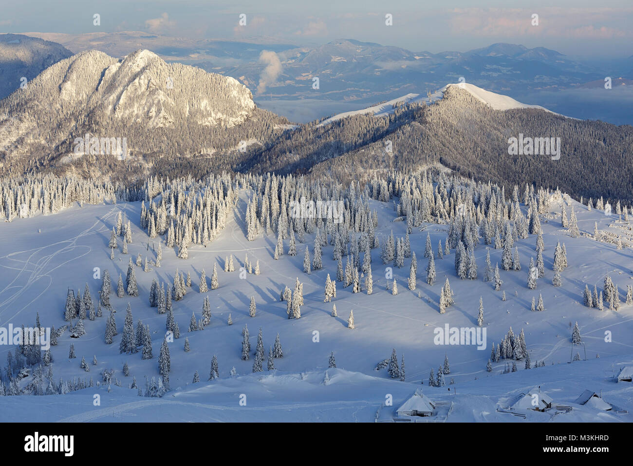 Szene im Winter auf Schnee bedeckten Plateau mit gefrorenen Pinien und Holzhütten, die Berge im Hintergrund. Stockfoto