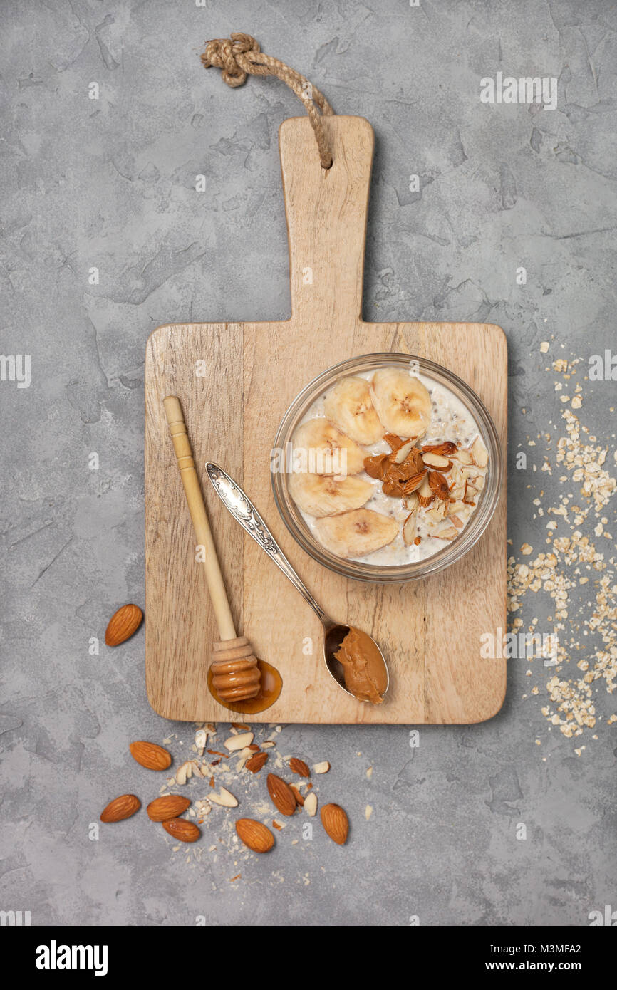 Gesunde Ernährung Frühstück. Übernachtung Hafermehl mit Chia Samen, Bananen, Peanut Butter, Honig in einem Glas auf einem grauen konkreten Hintergrund. Ansicht von oben Stockfoto