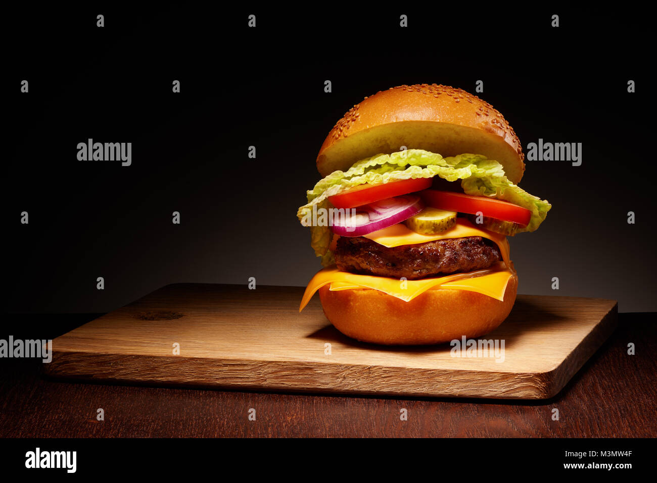 Lecker Hamburger mit Käse, Gurken, Tomaten, Zwiebeln und Salat auf Holz Schneidebrett auf dunklen Tabelle Stockfoto