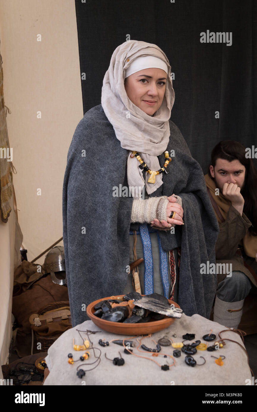 York, UK, 12. Februar 2018. Frau gekleidet wie ein Wikinger auf der jährlichen Jorvik Viking Festival. Sie ist die Teilnahme an einem realistischen Re-enactment von Leben bei einem Viking Markt & steht ein open-air abgewürgt. Auf dem Tisch liegt, ist eine Anzeige der Waren, die Sie verkaufen - Schmuck aus hauptsächlich von schwarzen Jet, sondern auch Bernstein und Fossilien. Ein Mann, der ist auch im Kostüm hinter ihr sitzen. Stadtzentrum von York, North Yorkshire, England, UK. Credit: Ian Lamond/Alamy Live Newscraft Stockfoto