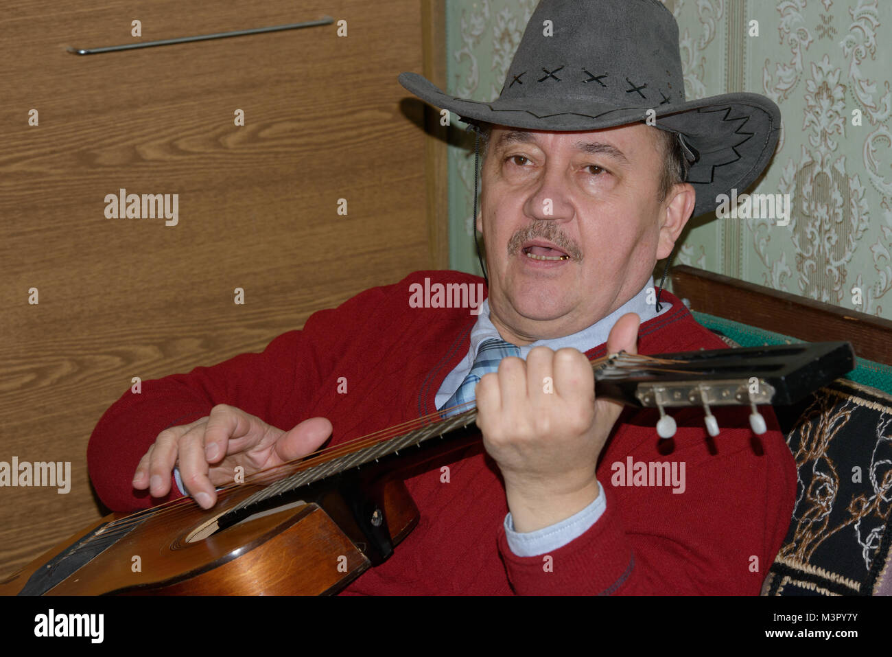 Kopf und Schulter Portrait von reifer Mann in grau Cowboyhut und kastanienbraunen Pullover, ist die Gitarre spielen und singen auf dem Sofa sitzt Home int Stockfoto