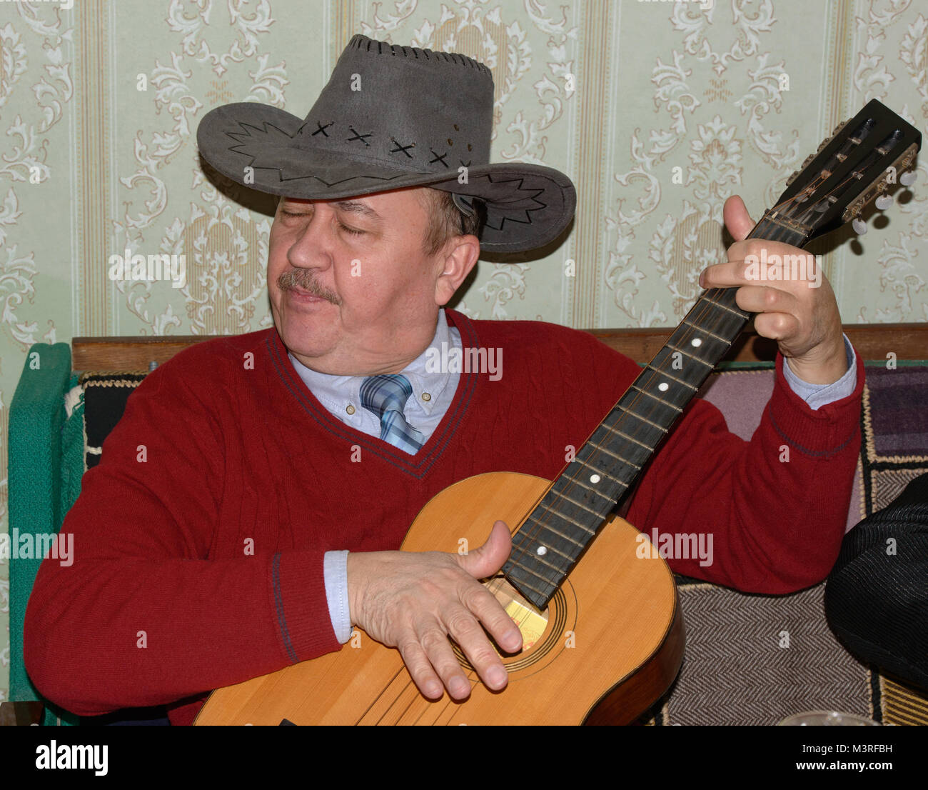 Kopf und Schulter Portrait von reifer Mann in grau Cowboyhut und kastanienbraunen Pullover, spielt die akustische Gitarre auf dem Sofa sitzt Home interi Stockfoto