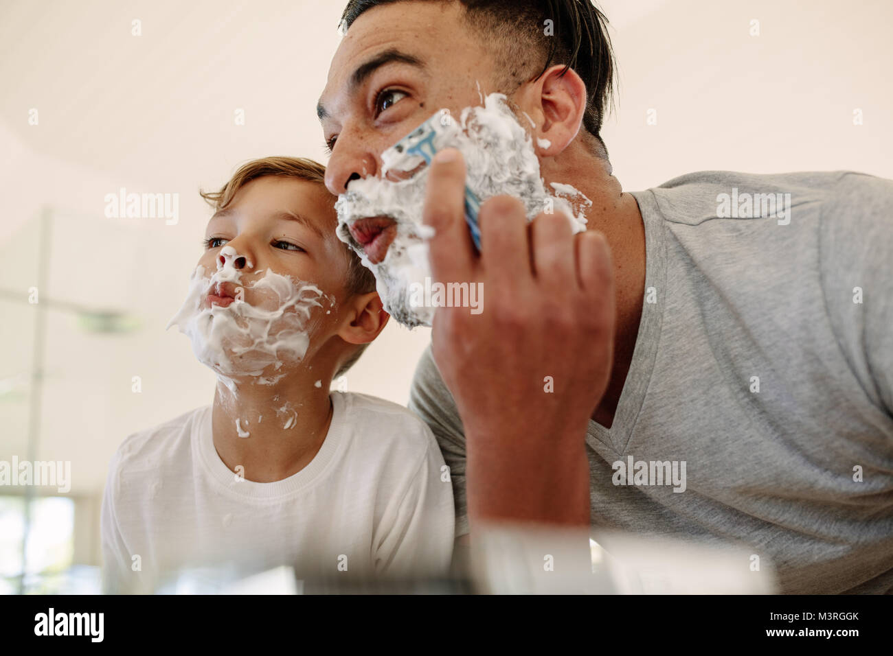 Vater und Sohn machen lustige Gesichter während der Rasur im Badezimmer. Junger Mann und der kleine Junge mit Rasierschaum im Gesicht rasieren, und Suchen in t Stockfoto
