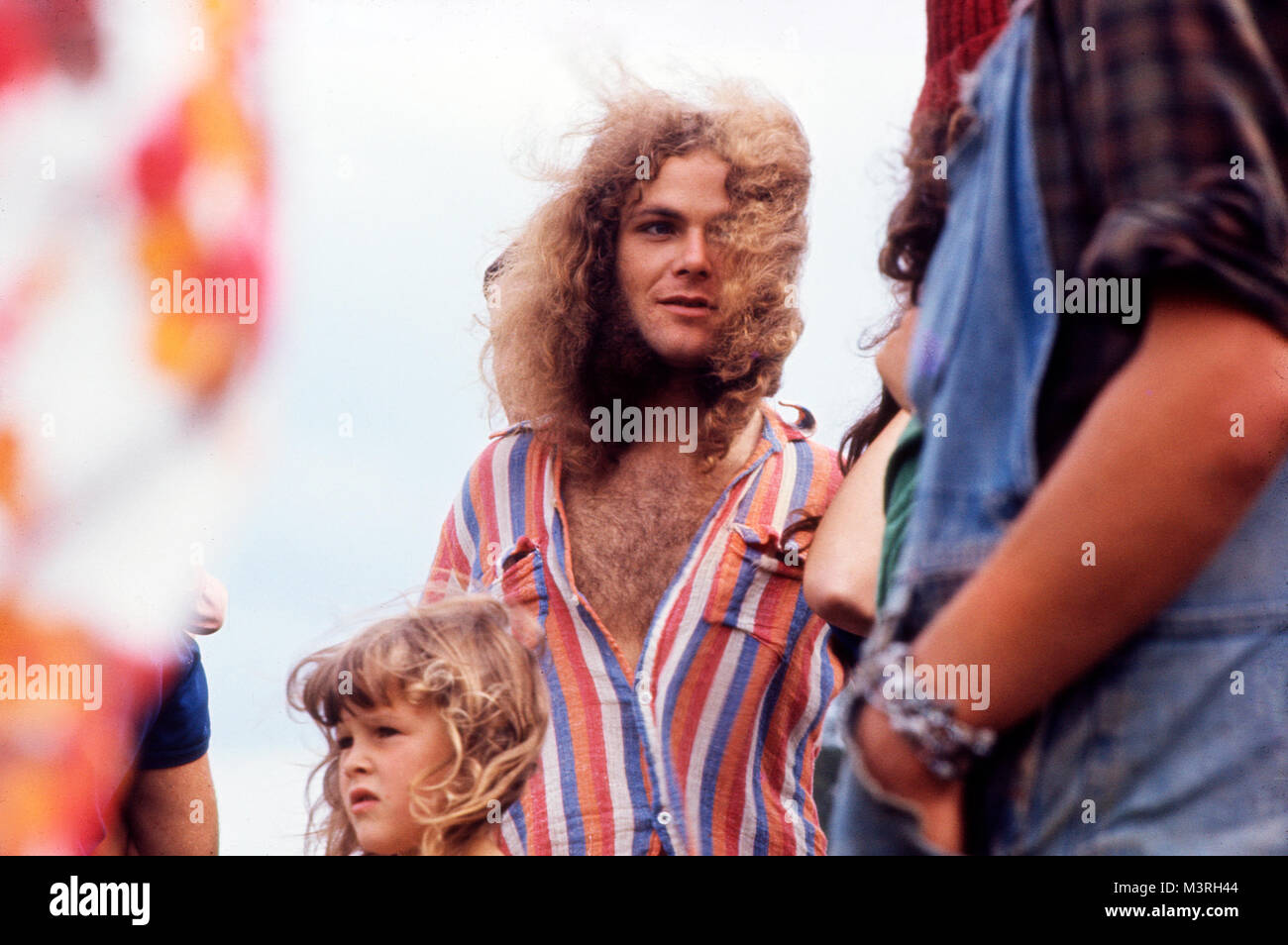 Haariger Mann mit langen Haaren, die im Wind wehen an einem windigen Tag mit einem Kind auf der Barsham Fair und Person, die Latzhose der 70er Jahre trägt, auf einem Musikfestival in Beccles Suffolk England Großbritannien 1974 KATHY DEWITT Stockfoto
