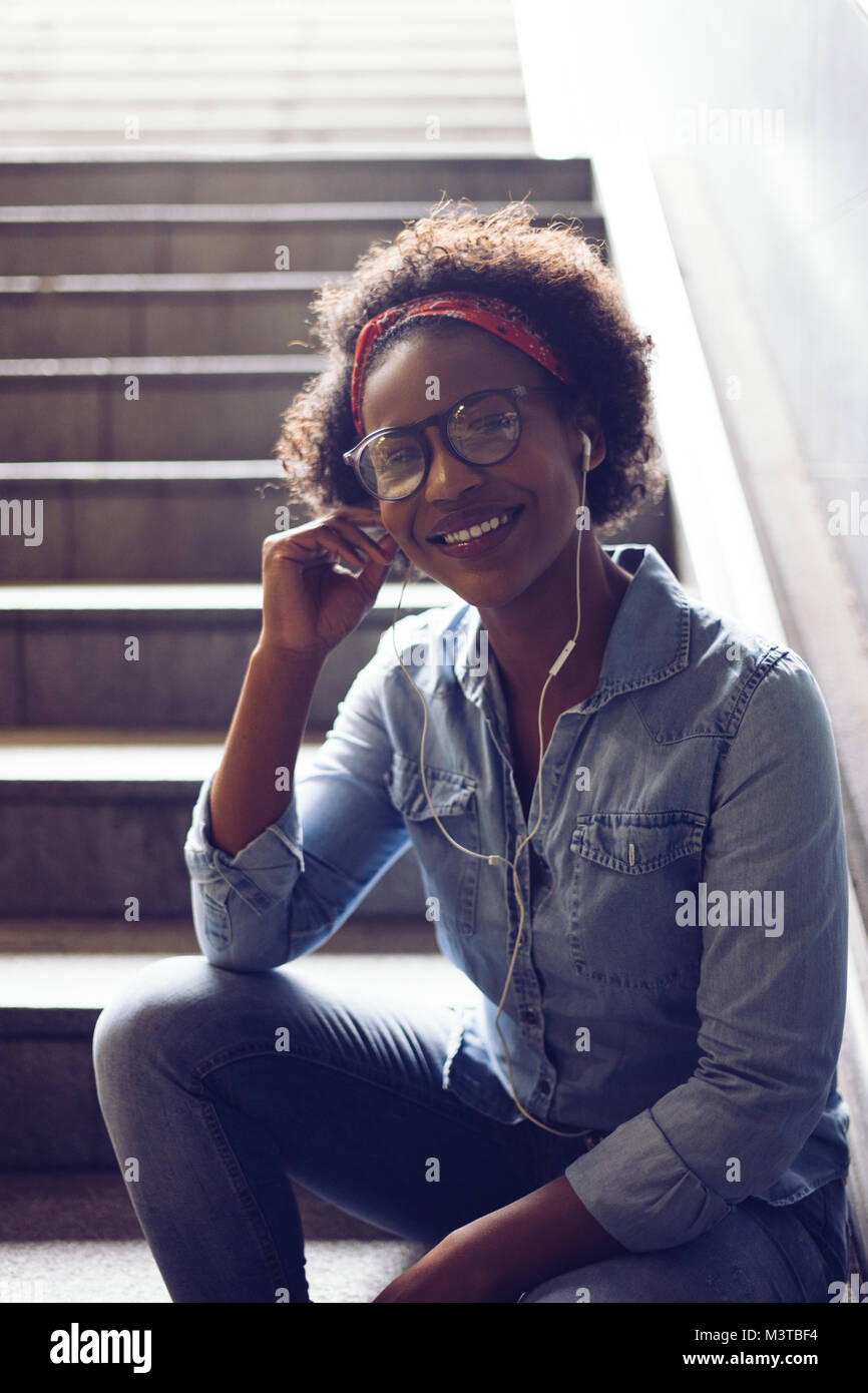 Stilvoll junge afrikanische Frau lächelnd, während allein an der Unterseite einer Treppe sitzen, Hören von Musik über Kopfhörer gekleidet Stockfoto