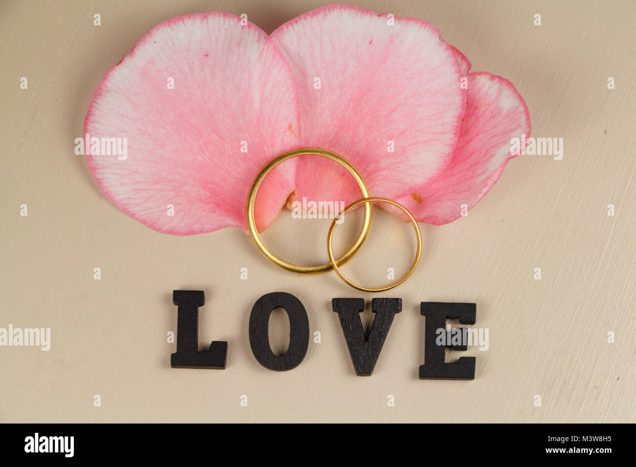Zwei Trauringe in Gold, drei rosa Blütenblätter der Blume und das Wort Liebe in schwarzen Buchstaben Stockfoto