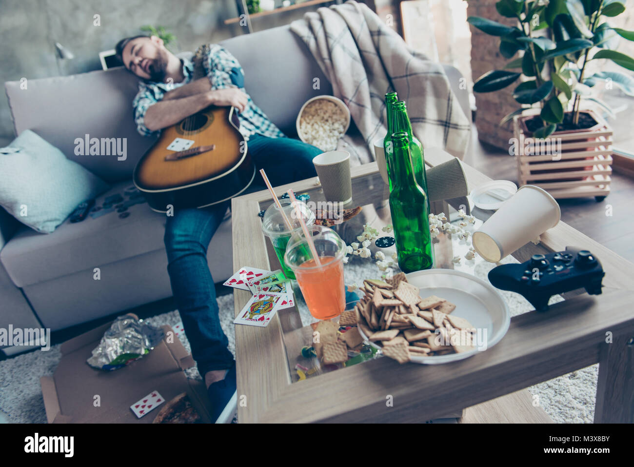 Nach der Party morgen, Bachelorette Party. Betrunken hipster umfasst eine akustische Gitarre schläft auf einem Sofa in unordentlichen voller Müll Zimmer Stockfoto