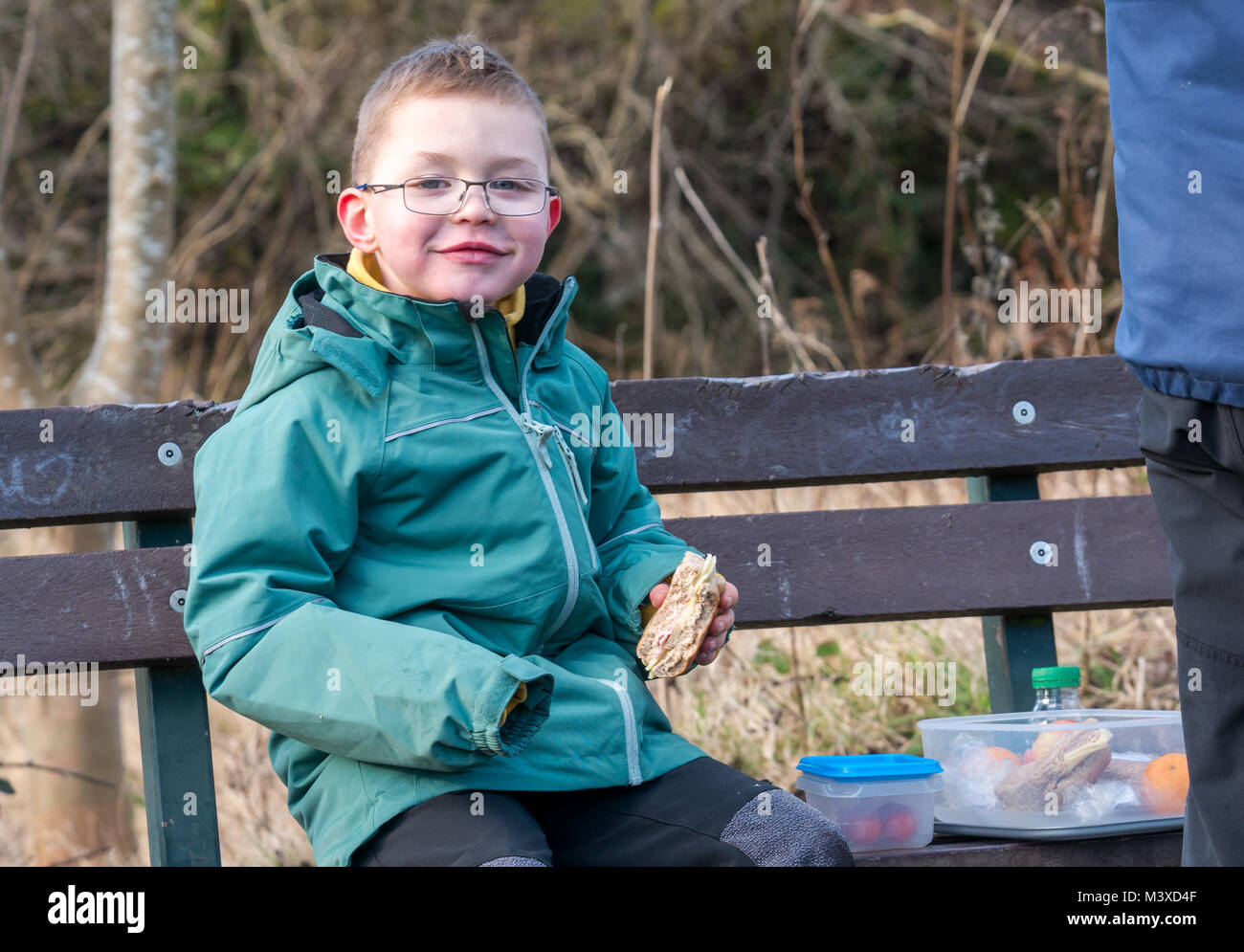 6 Jahr alten lächelnde junge Brille sitzen auf einer Bank bei einem Picknick Mittagessen. Er hält einen Schinken und Käse rollen, im Winter, Schottland, UK Stockfoto