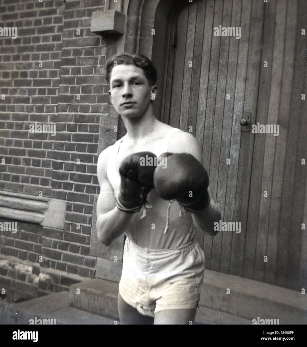 1950, historische... Die "edle Kunst' ... eine junge männliche Schüler außerhalb eines großen hölzernen Tür an Bancrofts Schule, Woodford Green, England, UK stehend, das Tragen eines Sport Weste und Hose und Boxhandschuhe und Stanzen Pose oder Haltung. Boxen ist ein Sport, der in den britischen Staat und Öffentlichkeit (privaten) Schulen in dieser Zeit gelehrt, und abgesehen von der Gesundheit und Fitness nutzen die Schülerinnen und Schüler erfahren, dass die Kämpfe in einem Ring statt, nicht auf der Straße. Stockfoto