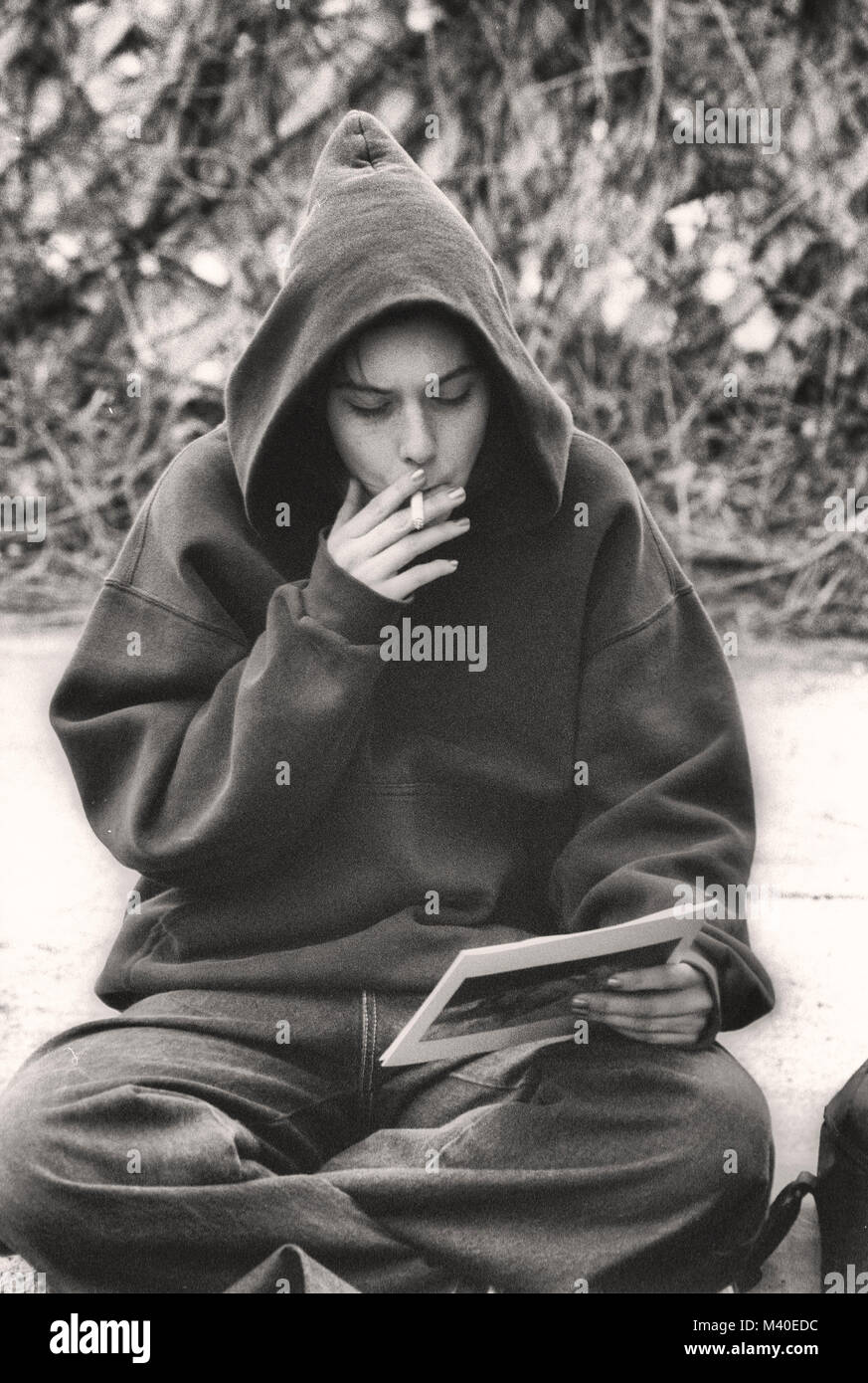 Ein junges Mädchen mit einem Hoodie, sitzend auf einer Kandare, rauchen eine Zigarette, mit Blick auf ein Foto. Stockfoto